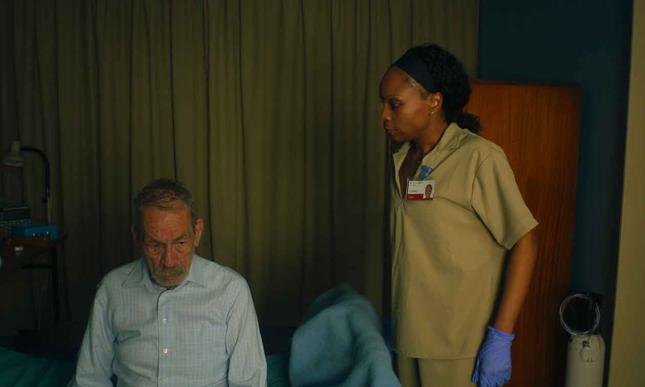 Still from the film: Arménio sitting on a care-home bed as the carer Hermínia stands by the door